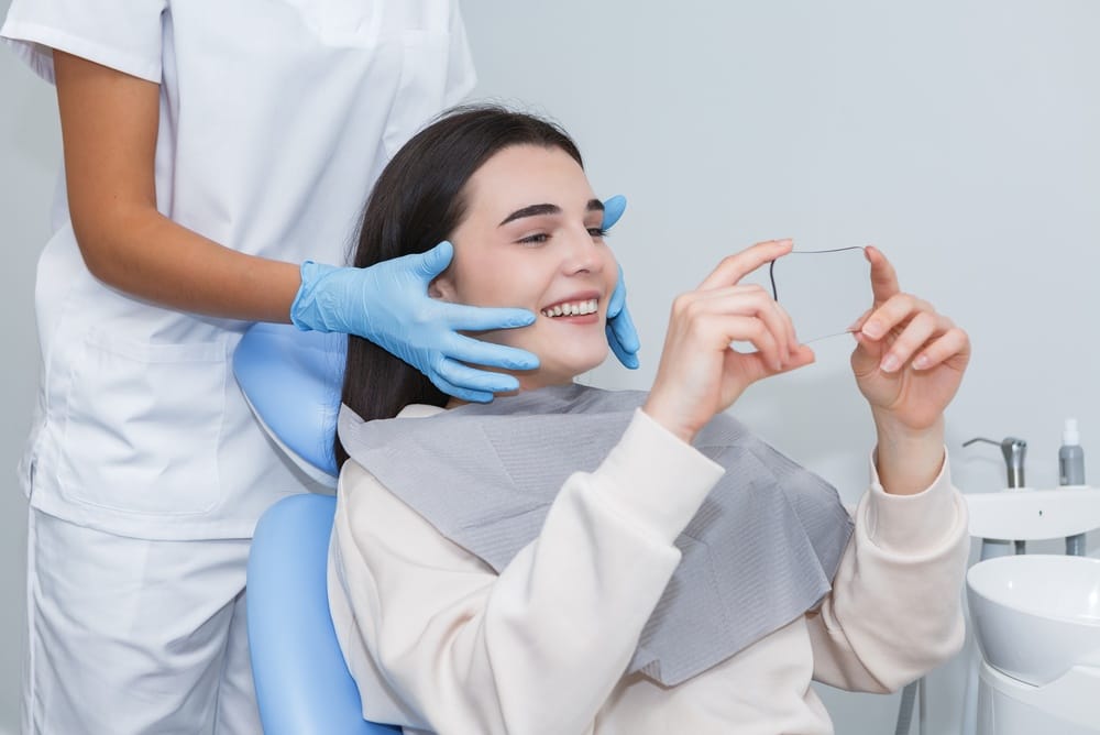A dentist is helping a patient take a selfie while sitting in a dental chair at a clinic. The patient is smiling with a dental bib around their neck.