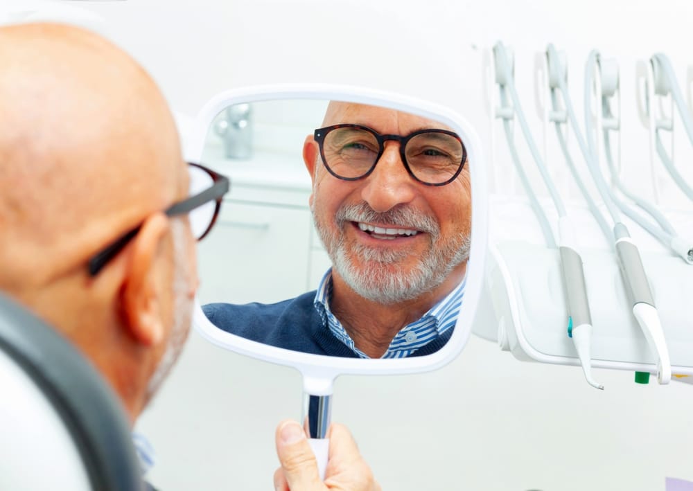 Happy senior man admiring his new smile in the small mirror at the dental clinic after a successful surgery