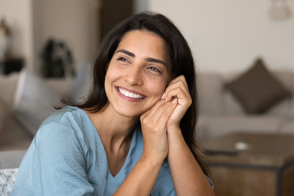 Joyful young Latin woman posing at cozy home, sitting in living room, leaning head and cheek on hands, looking away with toothy smile, opening healthy white teeth, thinking, dreaming