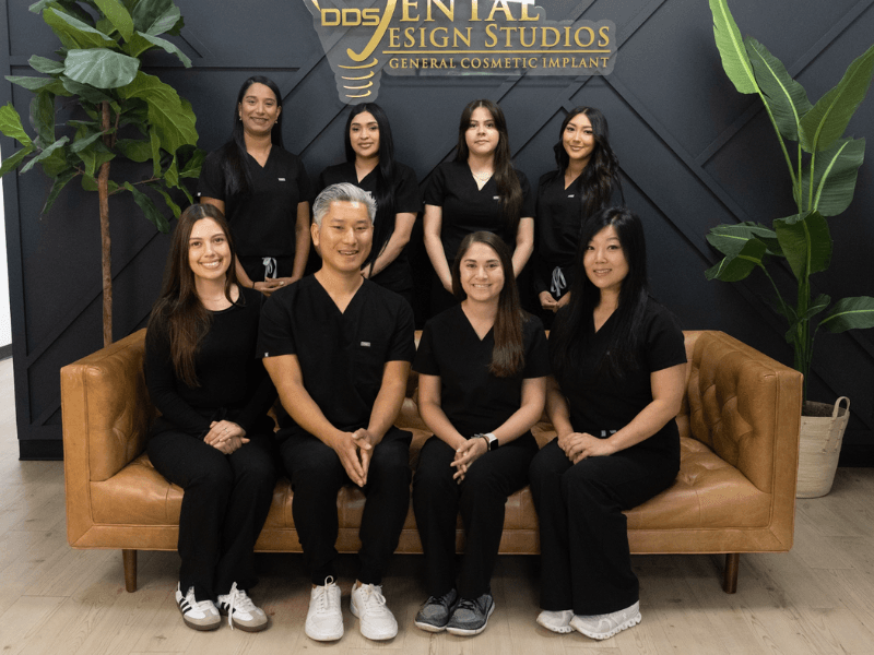 Dental Design Studios team group photo – Dental Design Studios LLC Dr. Andrew Kim, DDS, and the Dental Design Studios team posing together in the clinic lobby wearing black uniforms with the clinic logo in the background – Dental Design Studios LLC