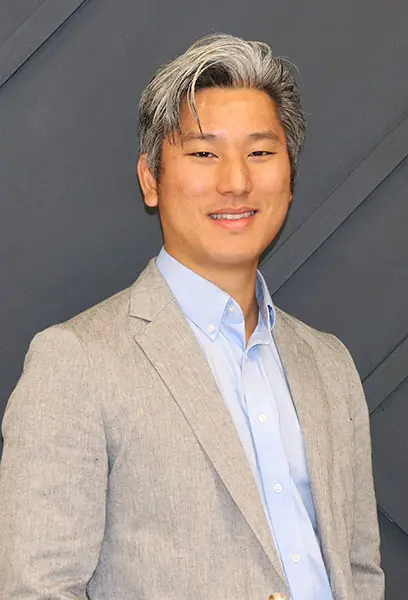 Dr. Andrew Kim, DDS smiling in professional attire – Dental Design Studios LLC Dr. Andrew Kim, DDS standing against a dark wall, wearing a light gray blazer and blue shirt, smiling confidently – Dental Design Studios LLC