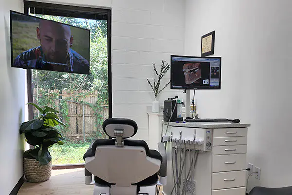 Dental treatment room with modern equipment and entertainment - Dental Design Studios LLC Well-lit dental operatory featuring a patient chair, mounted TV displaying a show, computer monitor with a dental scan, potted plants, and window view of a garden fence - Dental Design Studios LLC