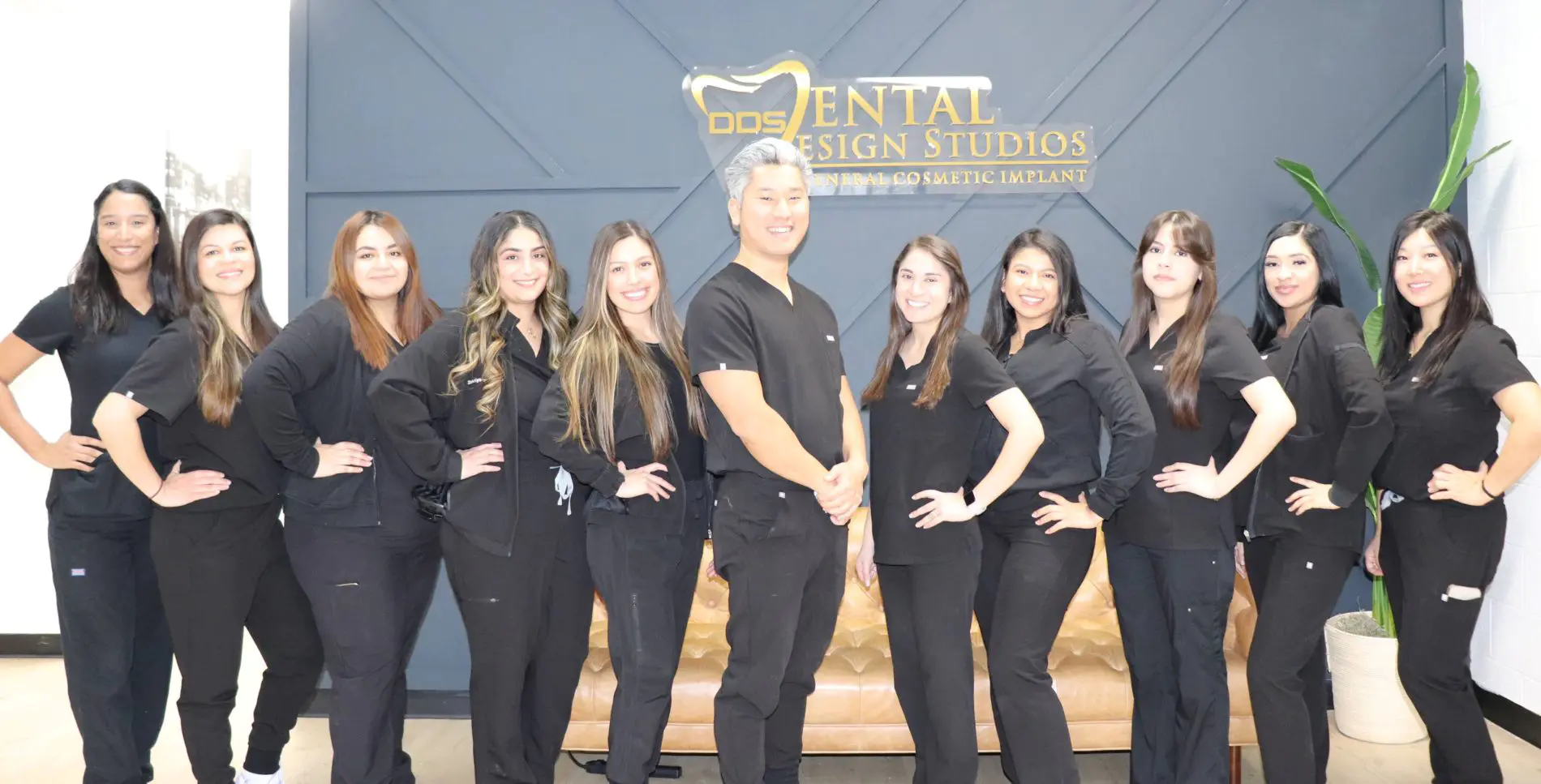 Dr. Andrew Kim, DDS, standing proudly with his all-female dental team in matching black scrubs, posing in front of the Dental Design Studios logo wall - Dental Design Studios LLC